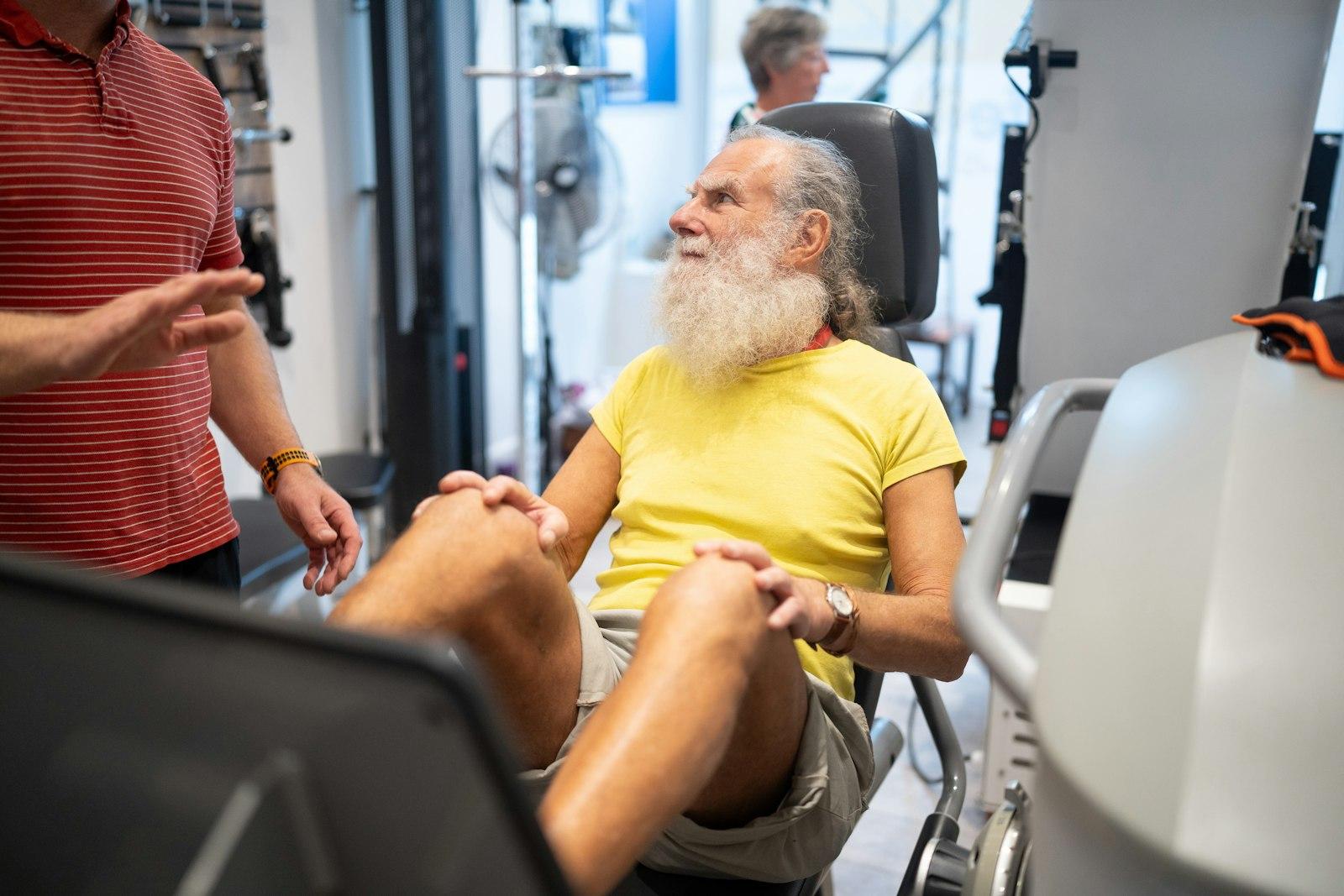 An older man in mid-conversation with a clinician beside him on a leg-press machine during a supervised gym-based rehab session