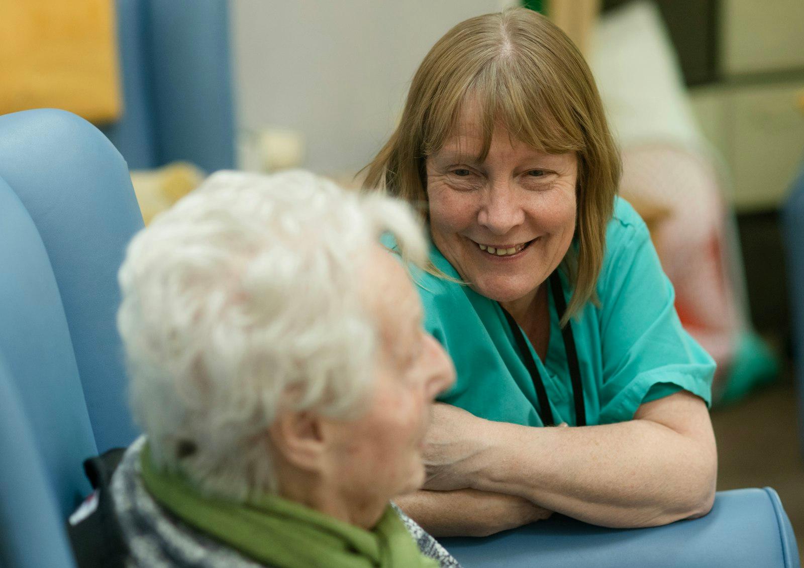 A clinician in scrubs leaning in warmly mid-conversation with an older woman seated in a high-back armchair