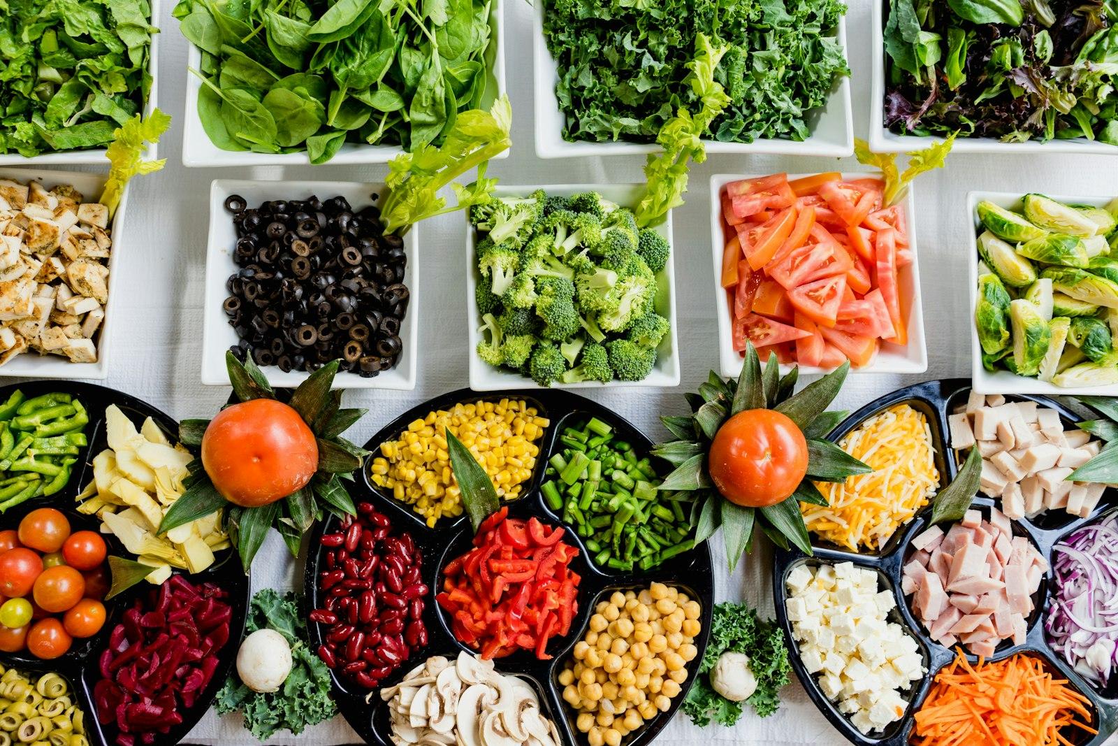 Top-down spread of fresh greens, broccoli, peppers, beans and tomatoes in white bowls