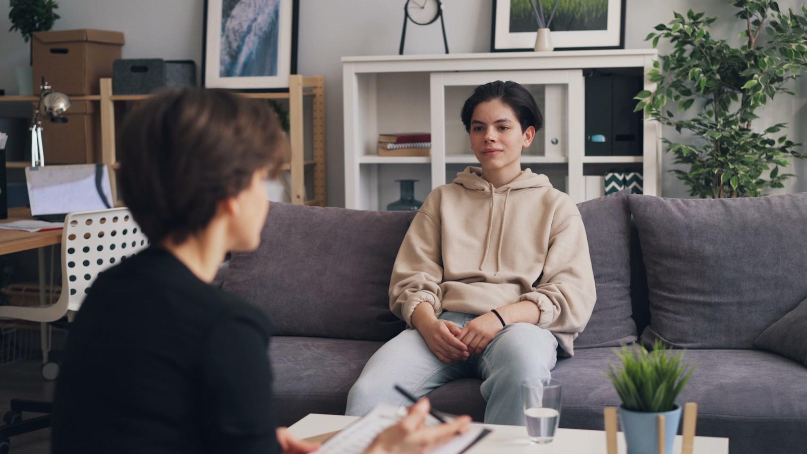 A teenage client and psychologist sit across from each other in a calm, warmly-lit therapy room