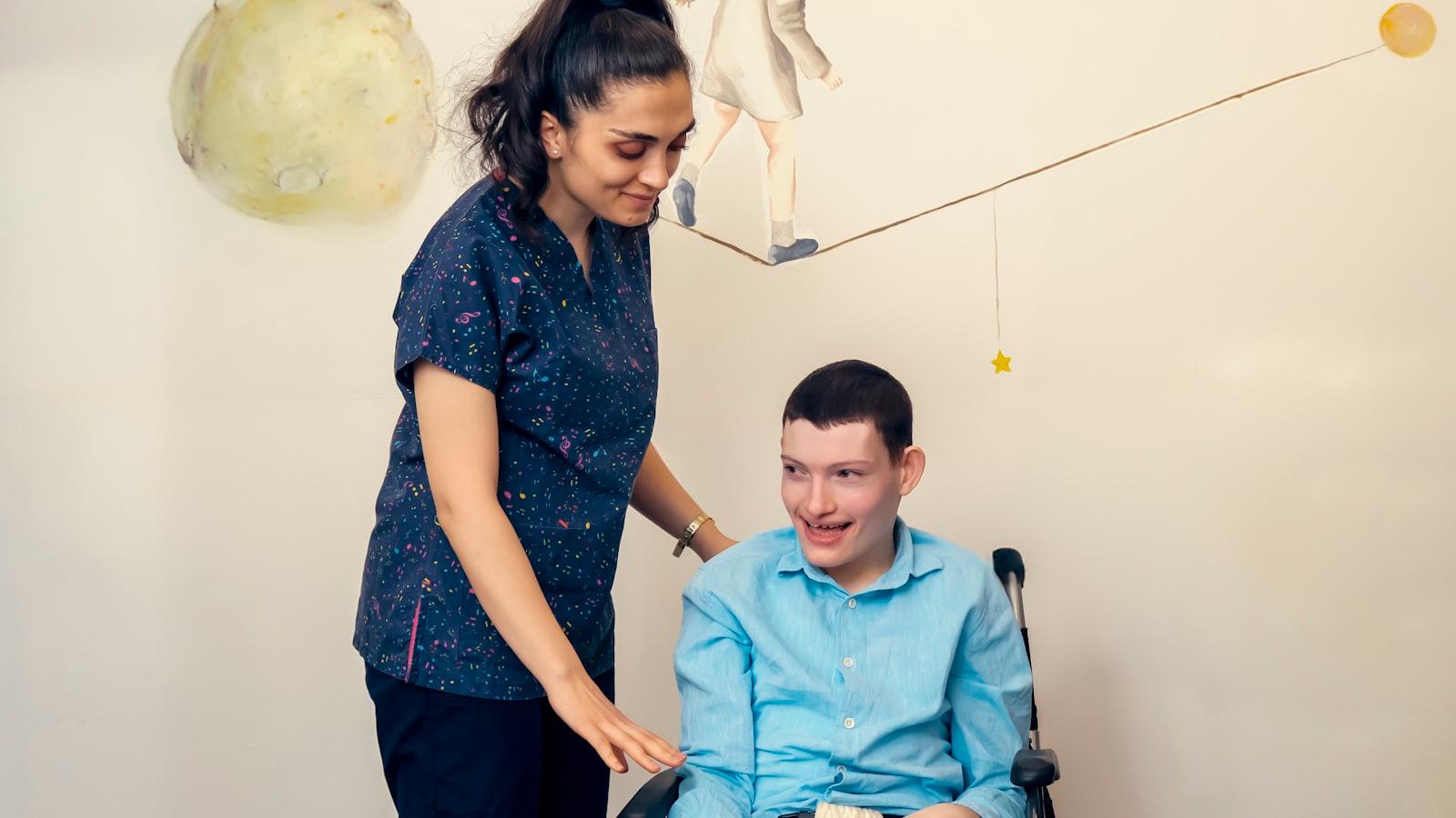 A clinician in scrubs leaning in warmly beside a young man seated in his wheelchair, in a therapy room with a hand-painted celestial mural