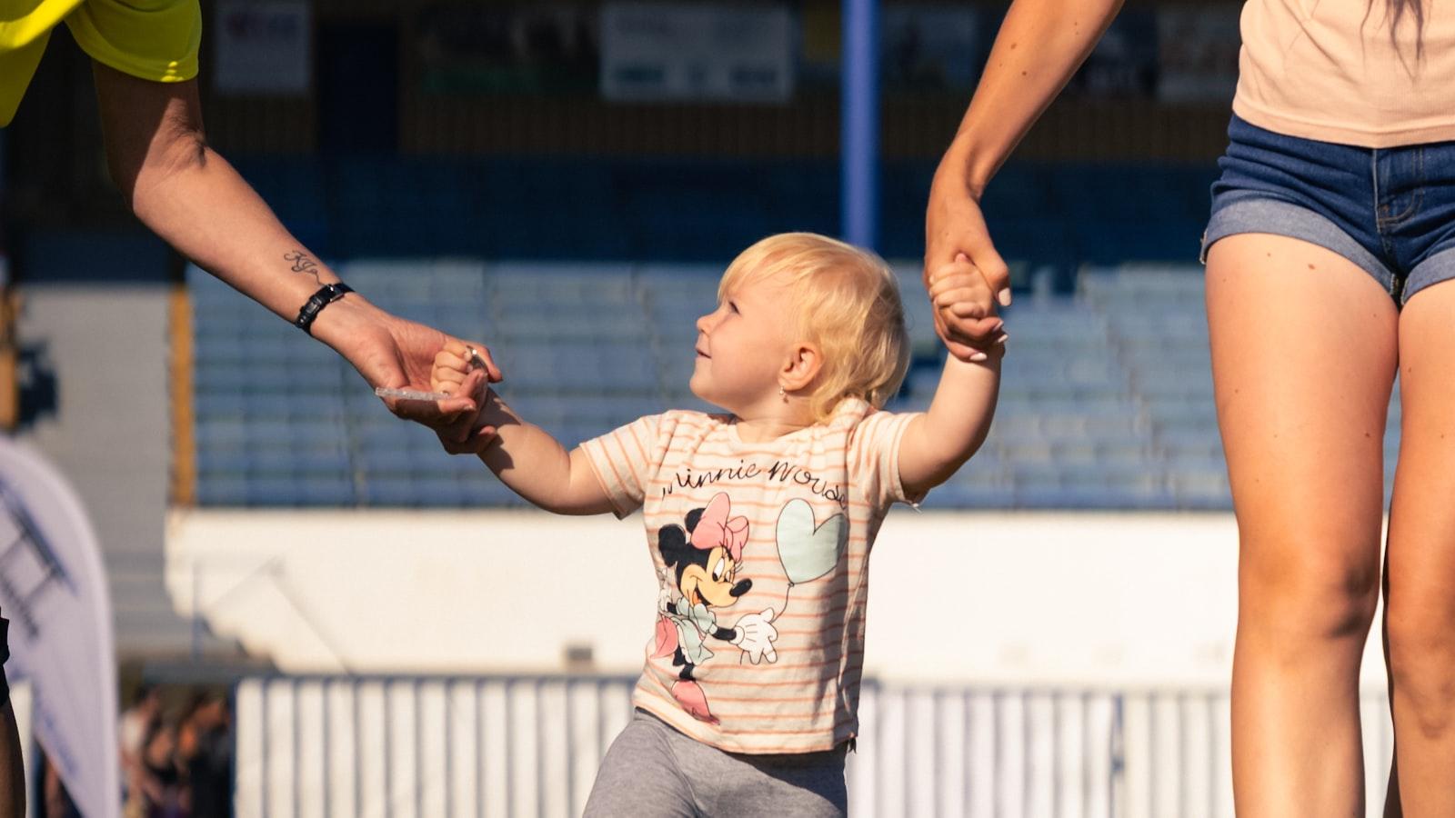 Toddler walking between two adults at sunset, gripping each of their hands as they cross a row of yellow agility hurdles