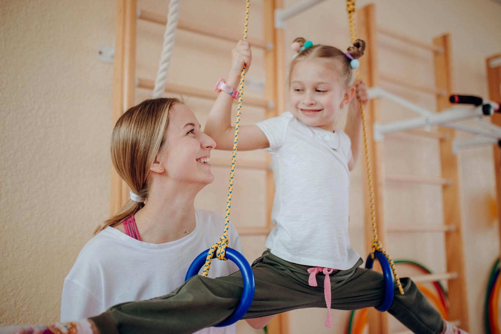 Girl with her legs hooked over gymnastic rings being spotted from below by a clinician in a paediatric therapy gym with wall bars, hoops and padded mats behind